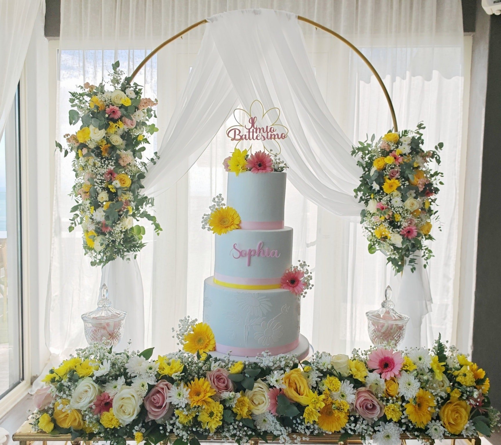 Allestimento battesimo con torta scenografica, fiori gialli e arco oro